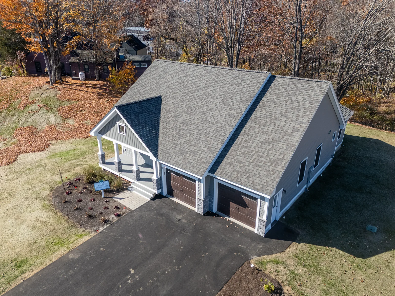 Aerial drone photo of a New England home with a gray shingle roof and double garage on a wooded autumn lot